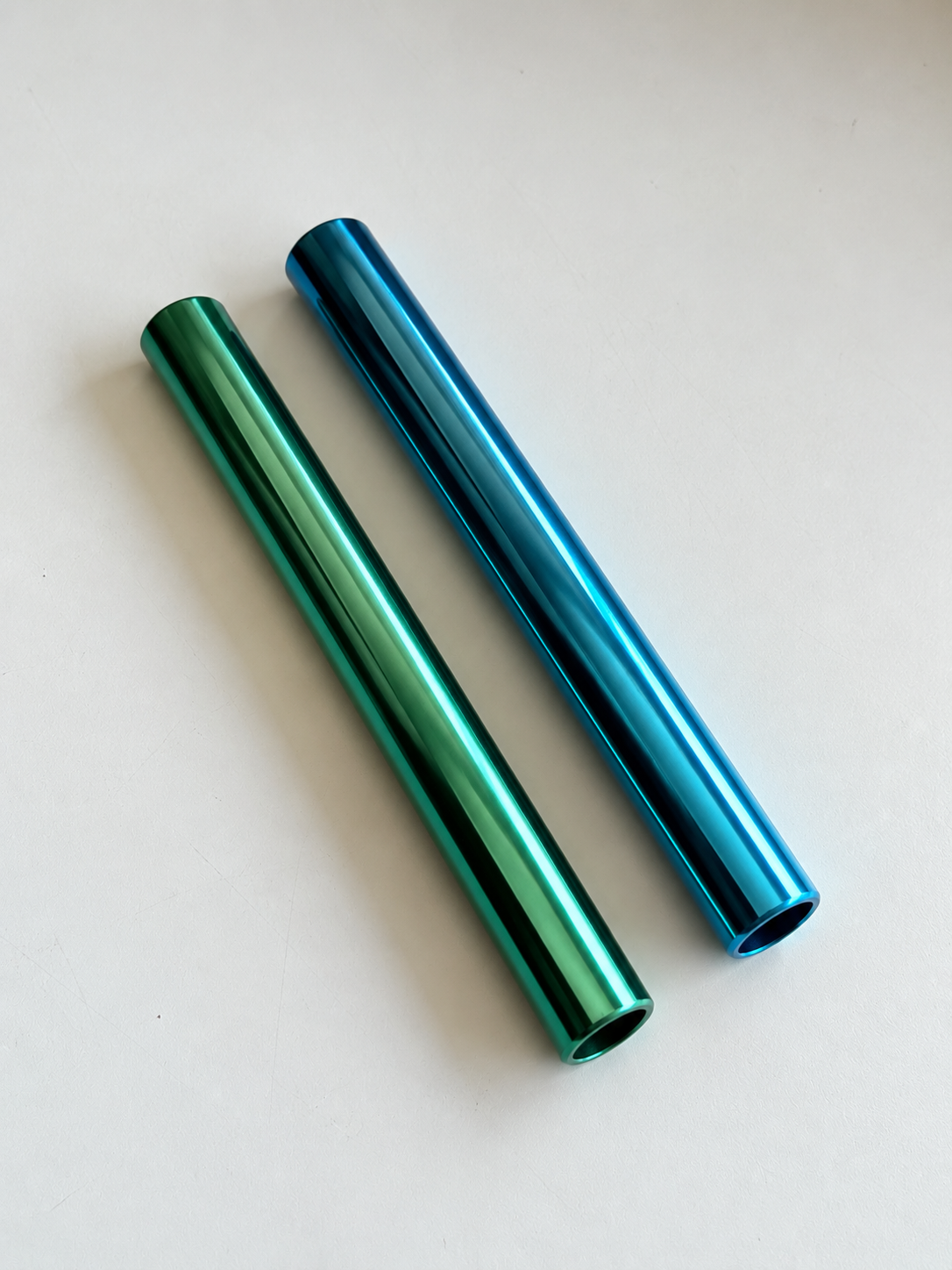 Grün-türkises und blaues glänzendes Aluminiumröhrchen, parallel und diagonal auf hellem Grund angeordnet.